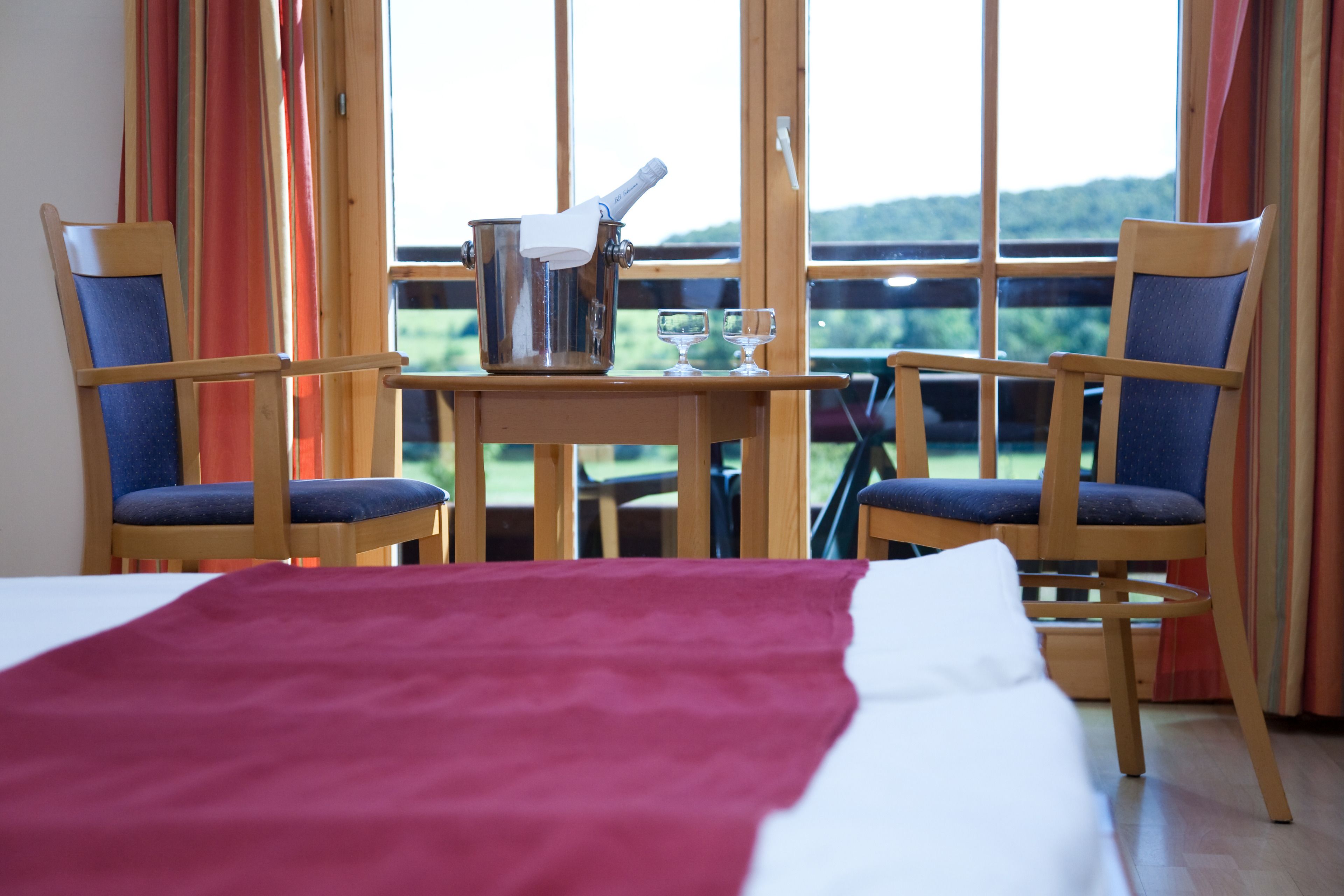 Une chambre d’hôtel avec un lit, deux chaises, une table avec une bouteille et des verres, et une vue sur l’extérieur.