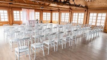 A large hall with wooden walls and ceiling, arranged with rows of white chairs and tables, large windows, and a chandelier.