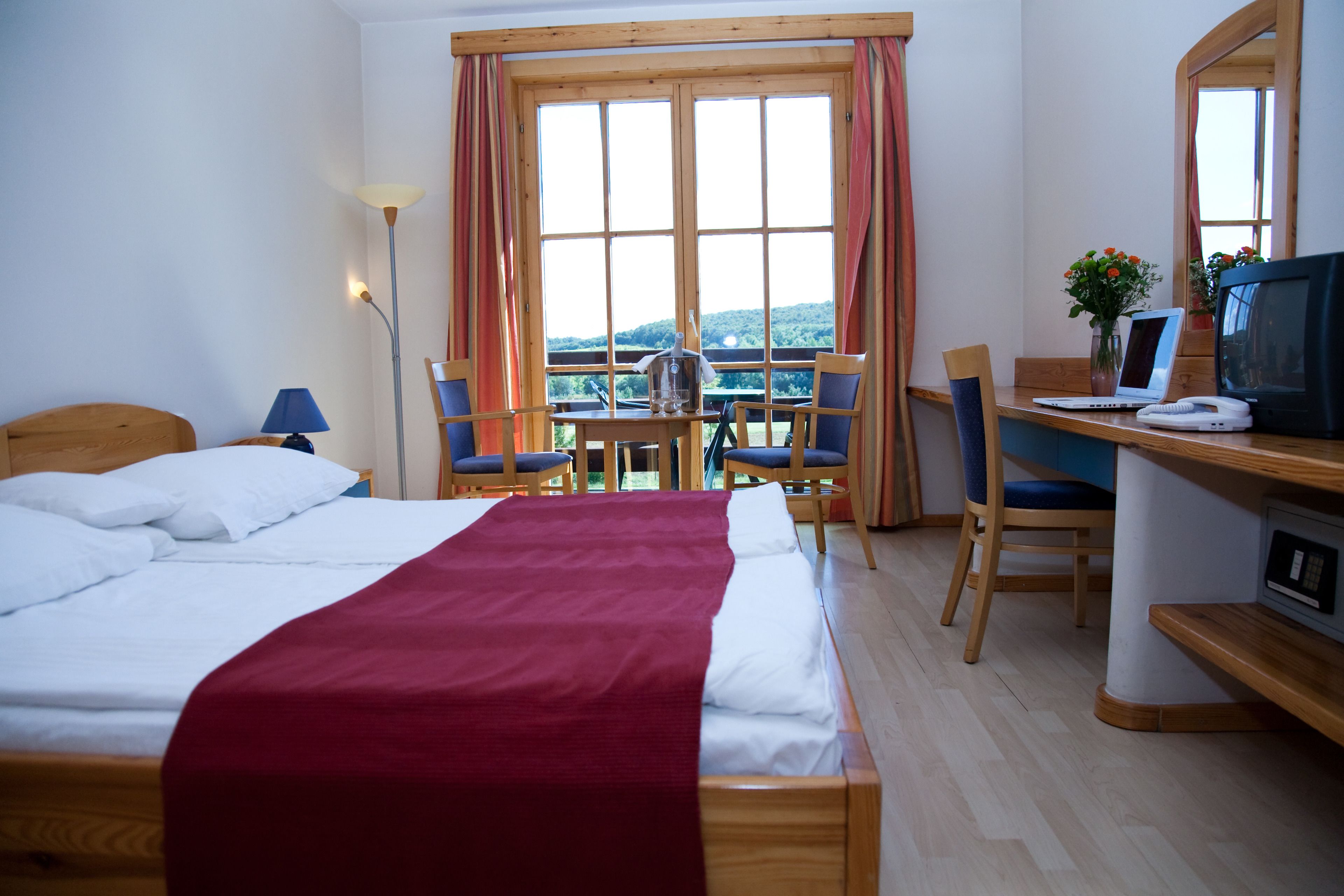 Ein Hotelzimmer mit Bett, Schreibtisch mit Computer, Stuhl, Lampe und Blick ins Freie.