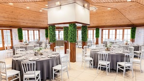 Um salão amplo com mesas redondas, cadeiras brancas e decorações com folhagem verde.