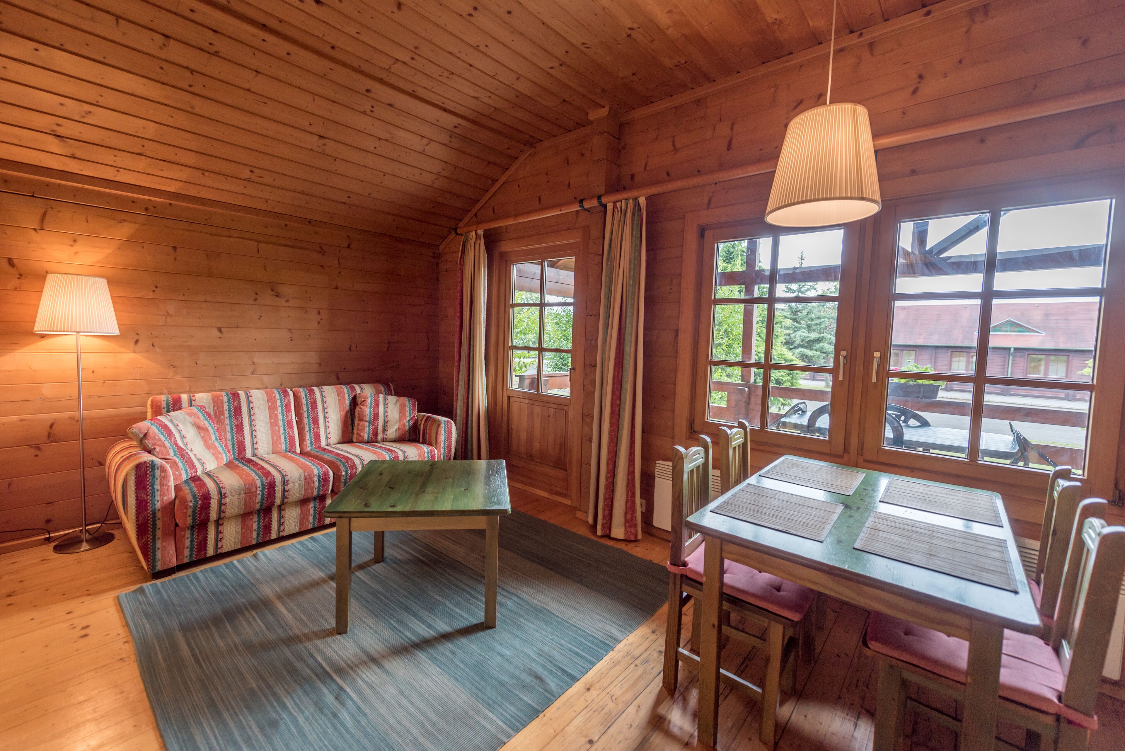 A cozy cabin interior with a sofa, table, and chairs, wooden walls, and large windows.