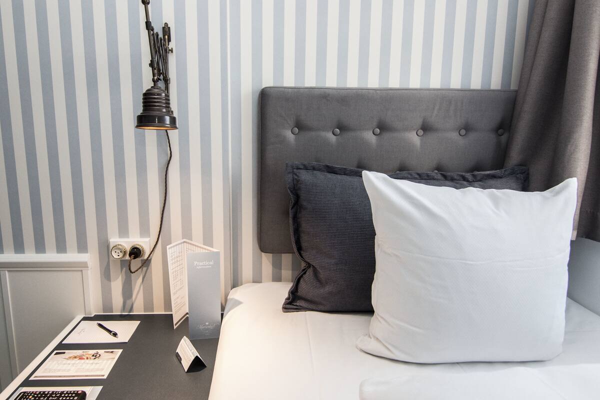 Una cama bien hecha con un cabecero gris, una almohada blanca y otra almohada gris oscuro. Una mesita de noche sostiene un control remoto, un bolígrafo y un folleto.