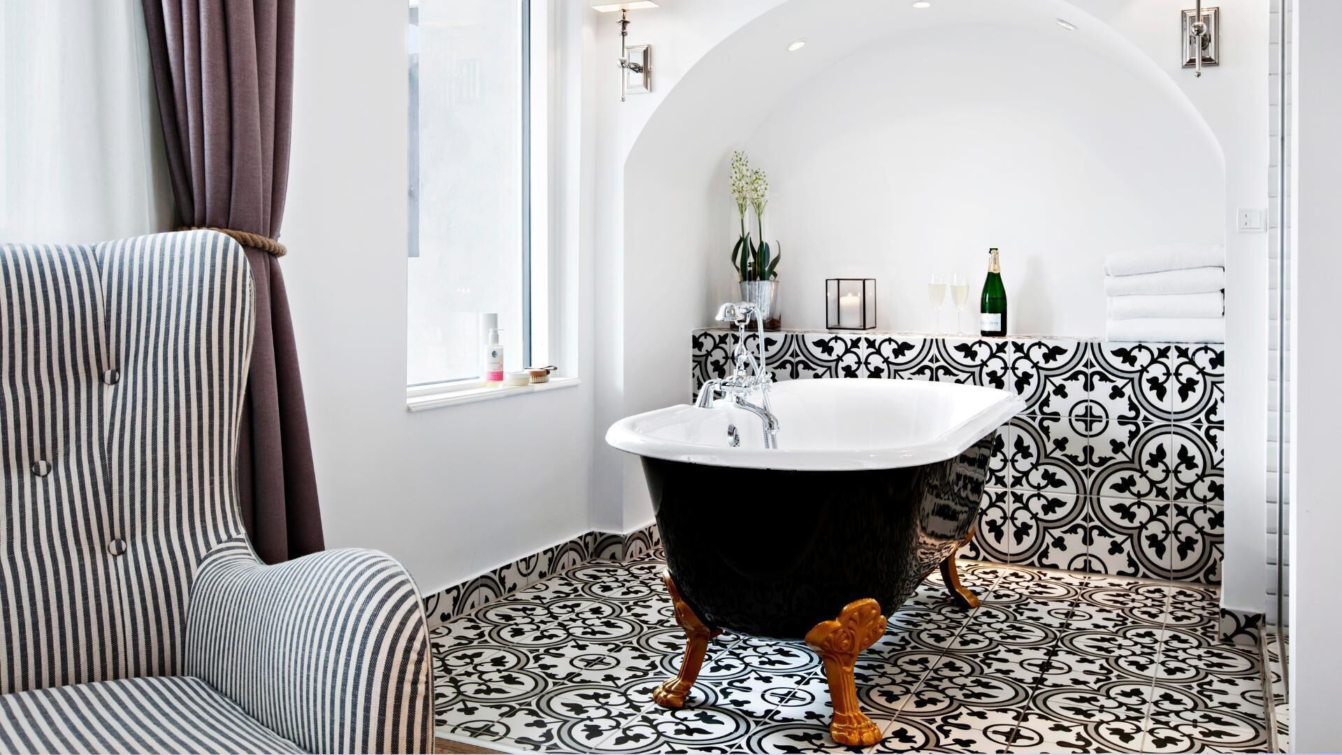 A bathroom with a black bathtub, striped chair, patterned floor tiles, and a window with curtains.