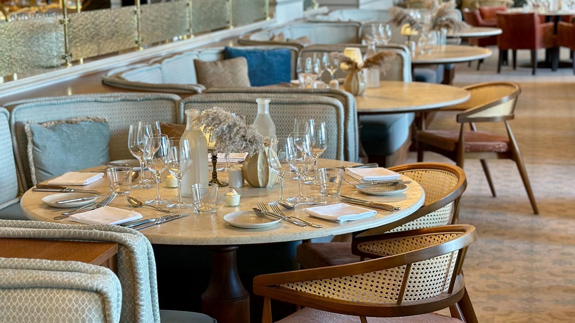 A restaurant with round tables set for dining, featuring a mix of upholstered chairs and wicker chairs.