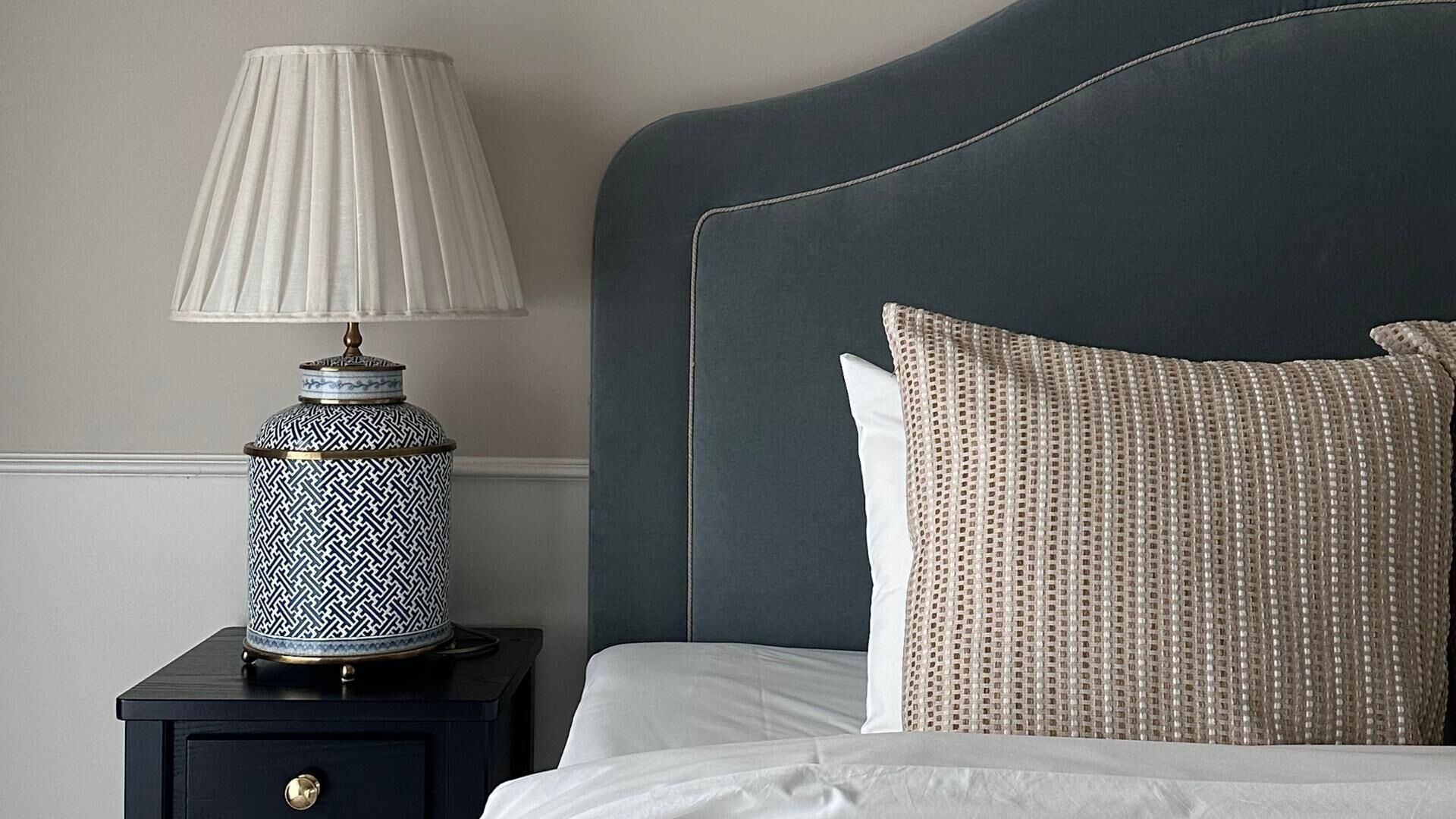 A neatly made bed with a dark headboard, a white pillow, and a patterned pillow. There is a small bedside table with a lamp and a decorative vase.