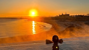 Une personne se trouve dans un bain à remous, d’où elle peut admirer un coucher de soleil sur une étendue d’eau gelée, avec un bâtiment visible au loin.