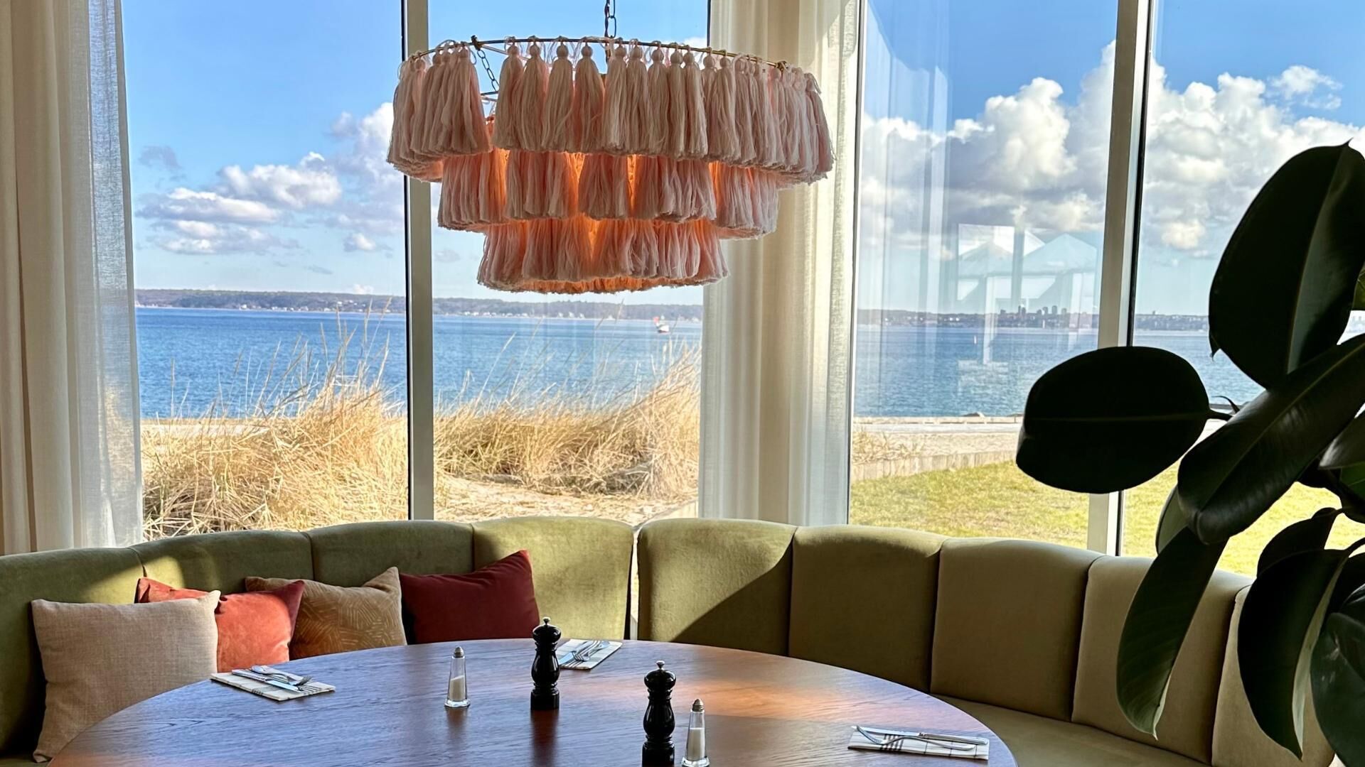 A room with a large window overlooking a body of water, a green cushioned seating area, and a wooden table with salt and pepper shakers.