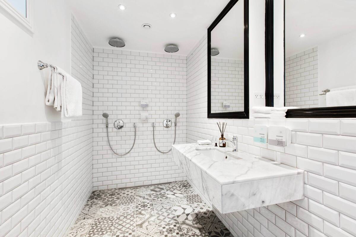 Un baño moderno con azulejos blancos tipo metro, piso estampado, tocador blanco con espejo y dos cabezales de ducha.