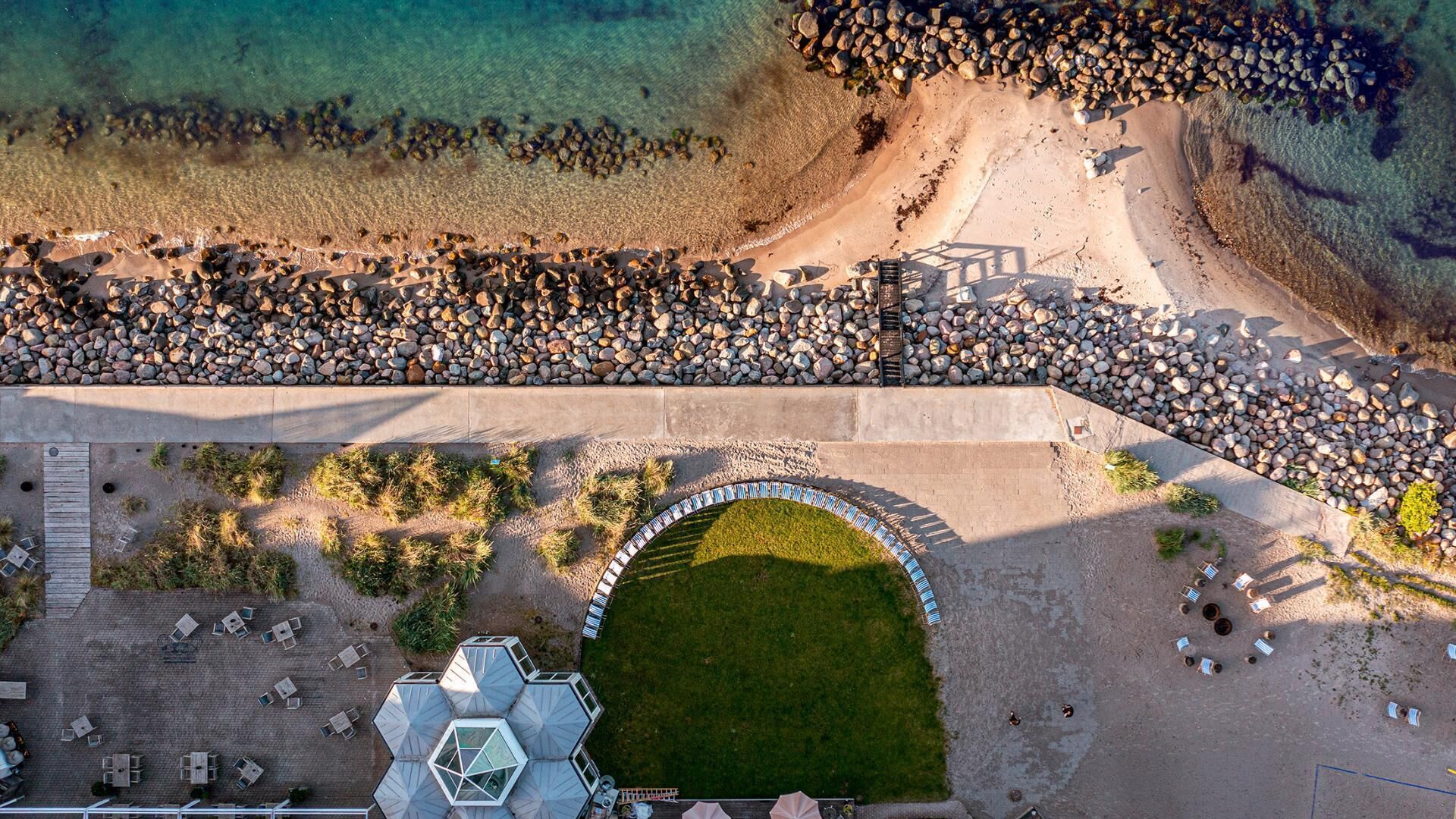 Vue aérienne d’un complexe hôtelier en bord de mer, avec une grande piscine, un espace de détente et une côte rocheuse.
