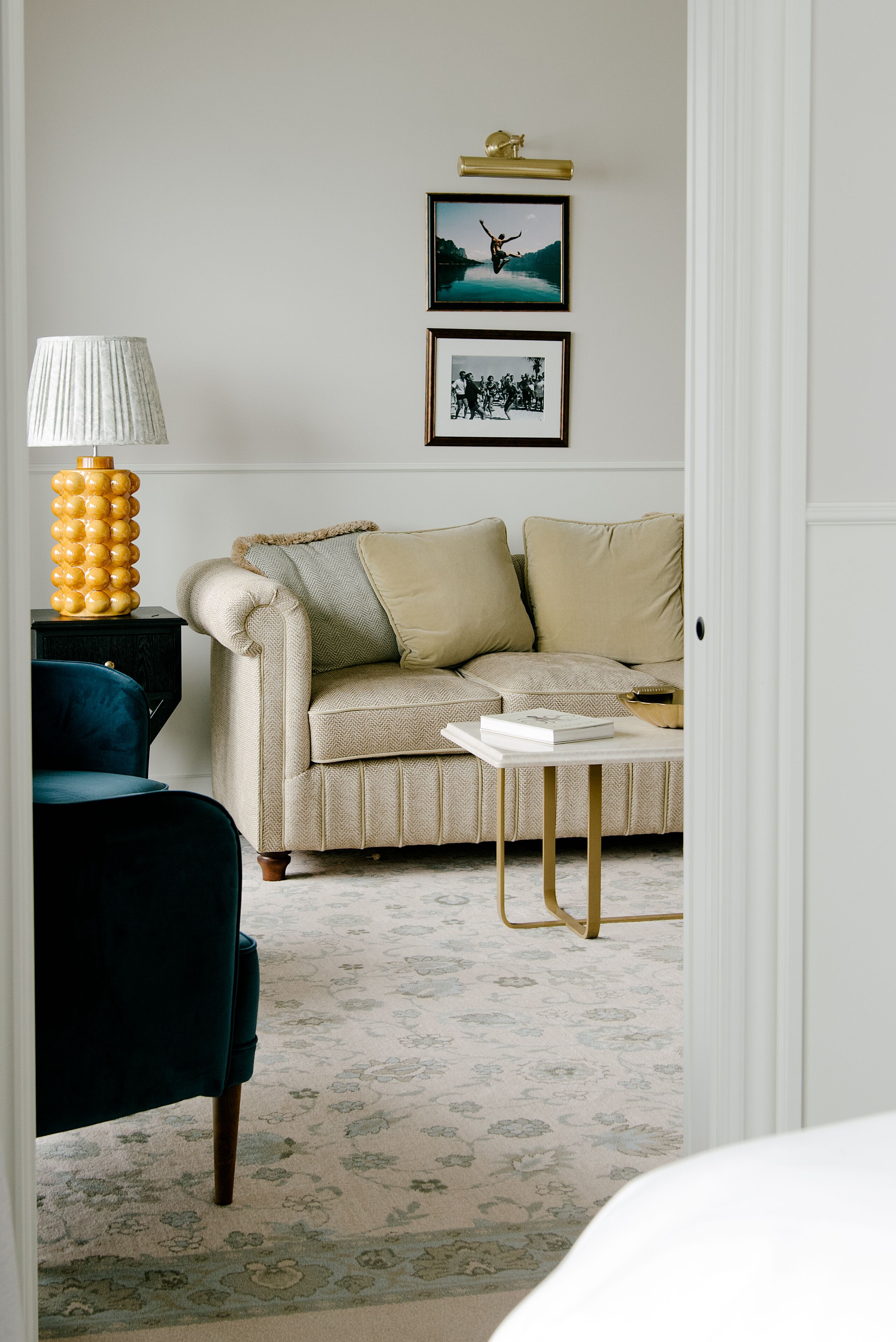 Un salón con un sofá color beige, un sillón azul oscuro, una mesita con un libro y una lámpara.