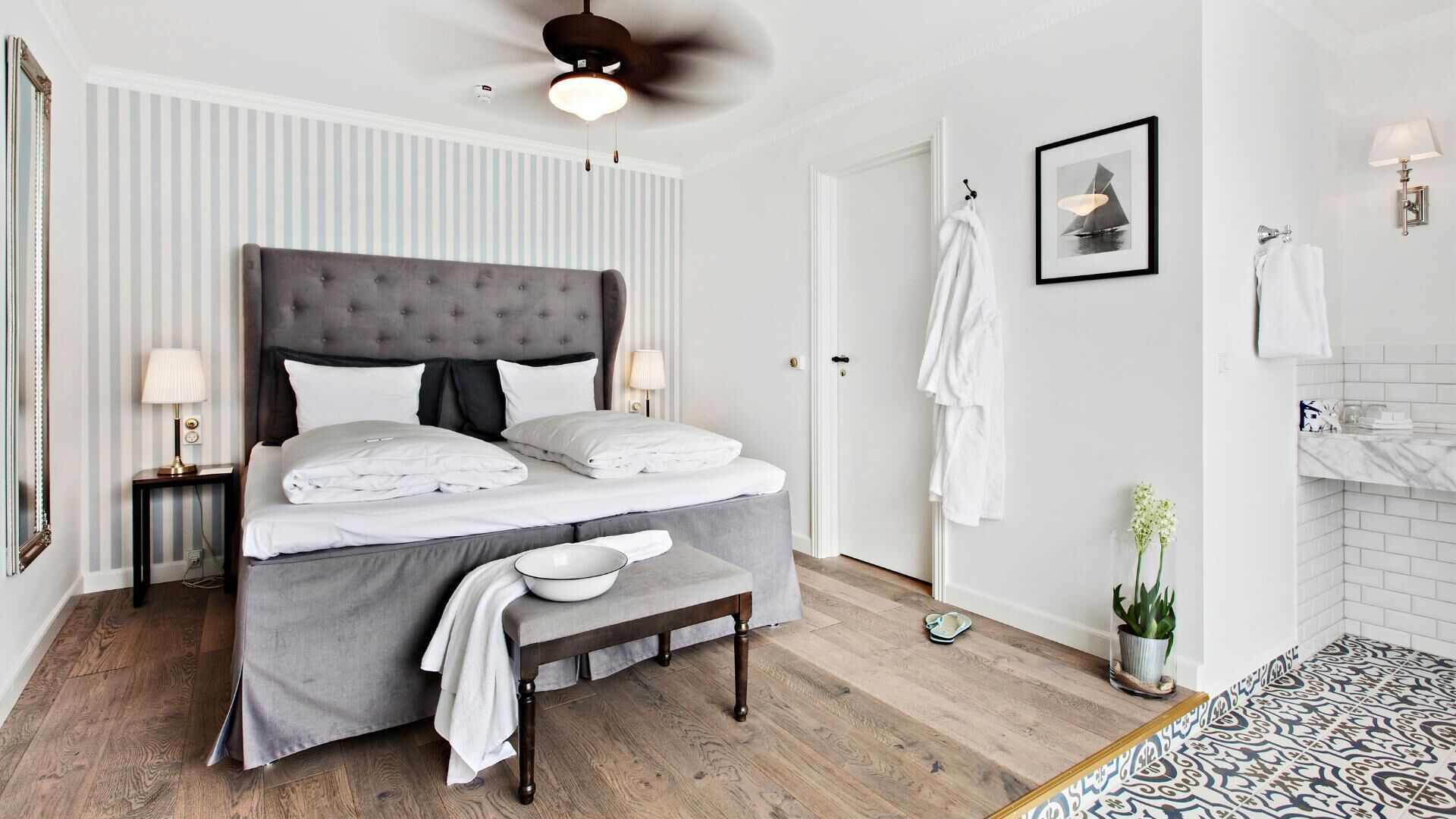 A modern bedroom with a tufted headboard, a bedside table with lamps, a bench, and a view of the bathroom.
