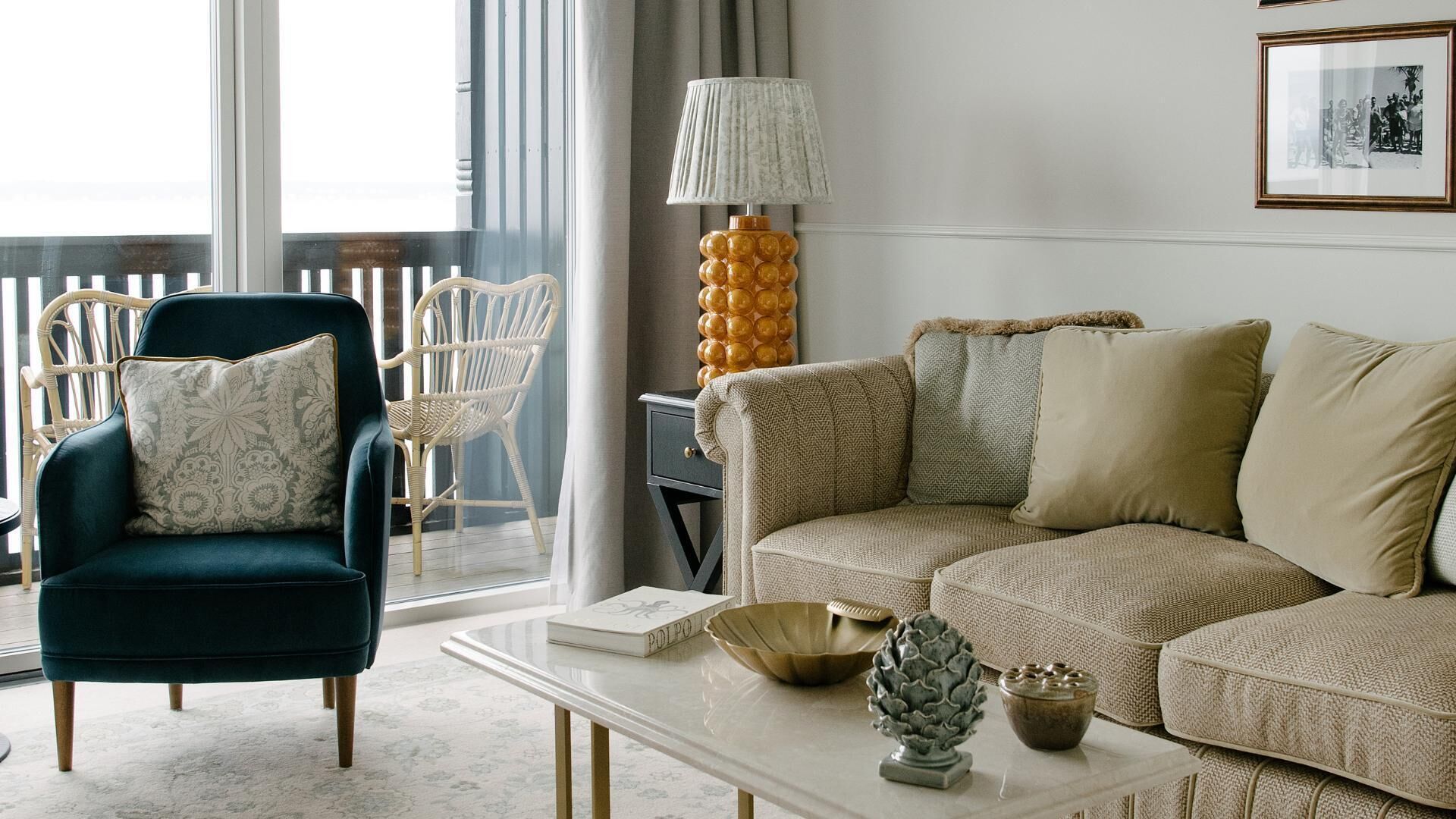 A living room with a teal armchair, a beige sofa, a white coffee table, and a lamp with a textured shade.