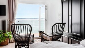 Une chambre d’hôtel avec un lit, deux fauteuils en osier, une télévision, une vue sur la mer et une fenêtre avec des rideaux.