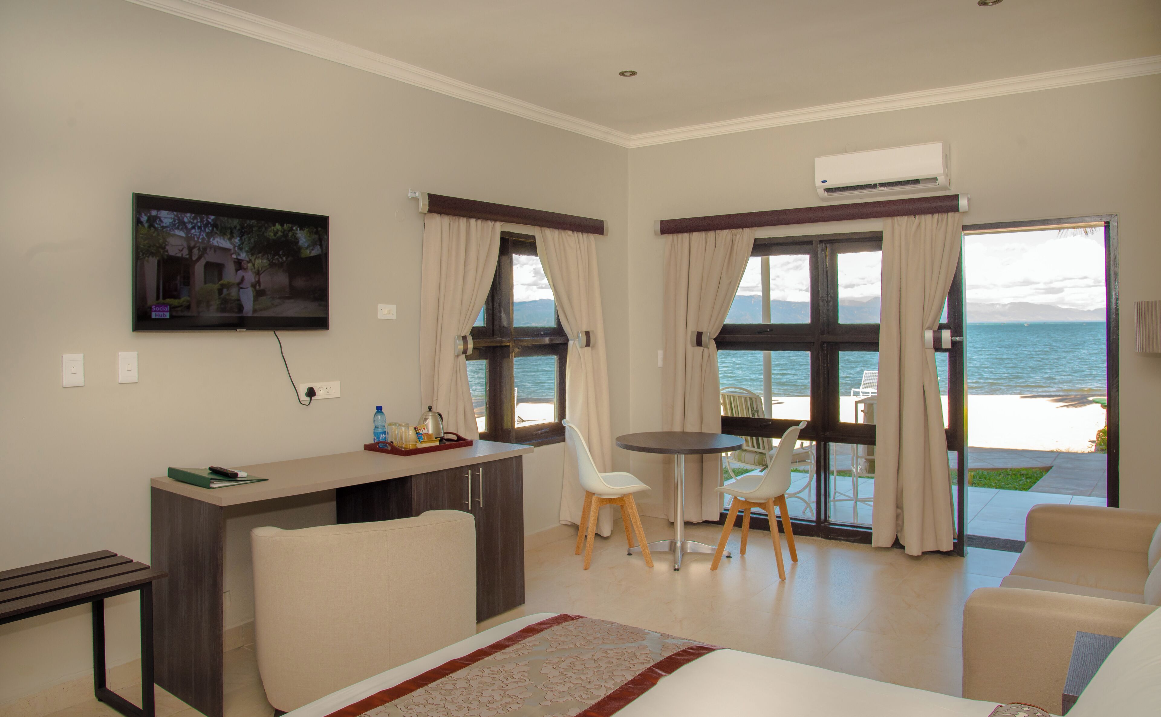 A hotel room with a bed, a desk, a chair, a small table, a TV, and a view of the sea.