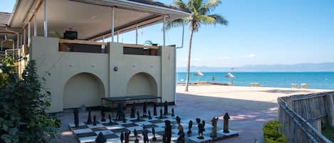 A beachside chess set on a checkered board, a covered seating area, and a palm tree.