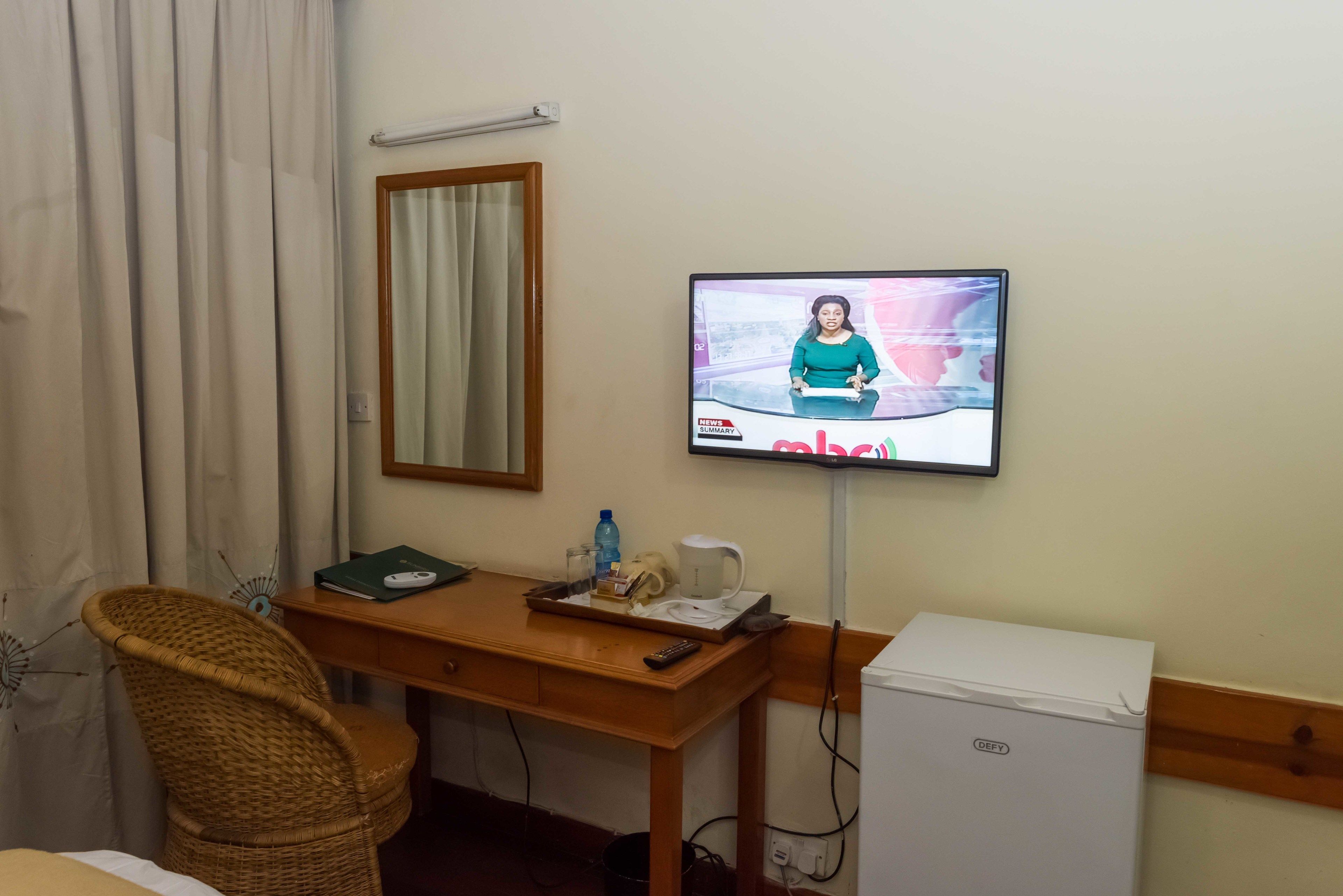 Habitación de hotel con escritorio de madera, una silla de mimbre, televisor montado en la pared, refrigerador blanco pequeño y ventana con cortinas.