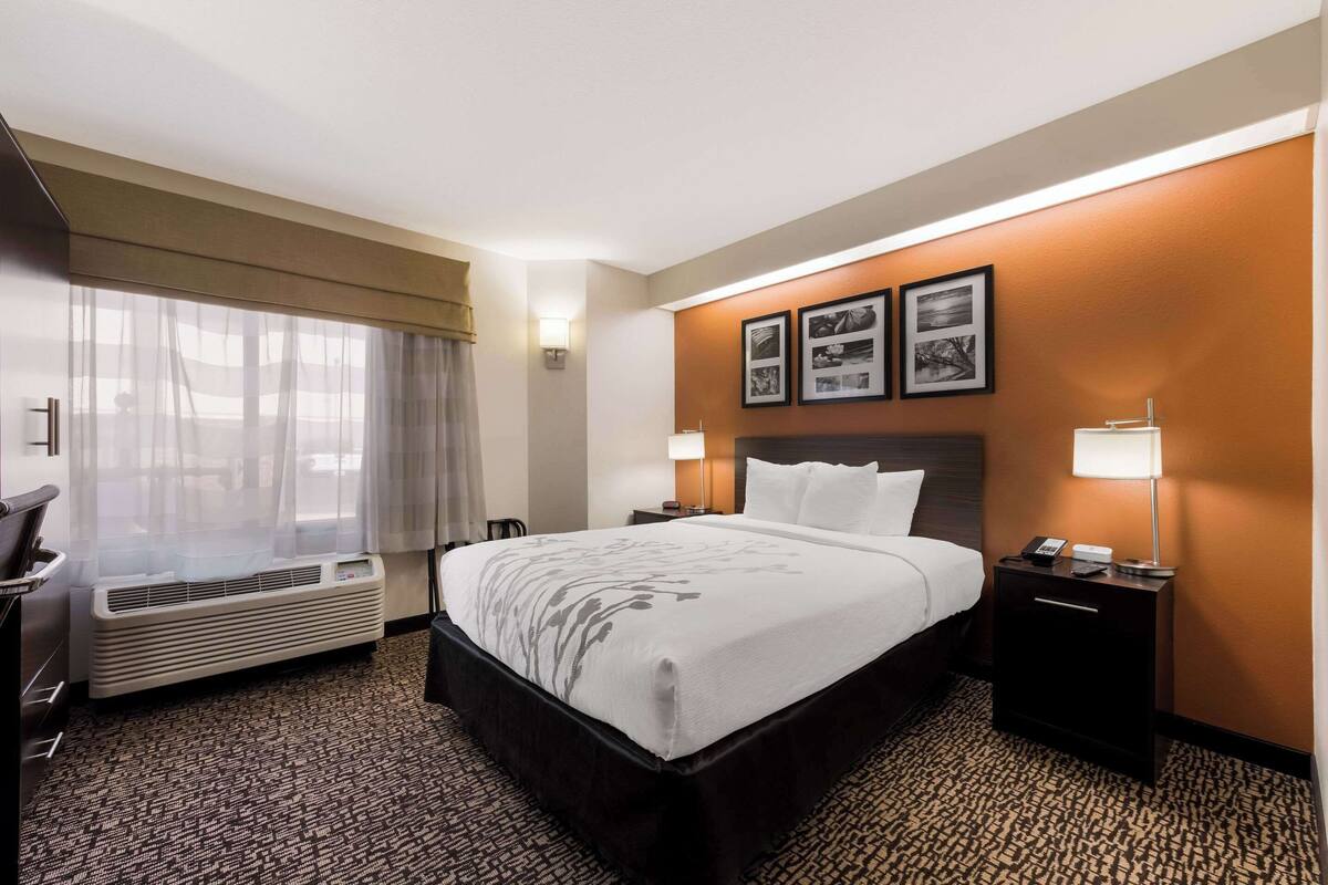 A hotel room with a large bed, a desk, a chair, an air conditioning unit, and framed pictures on the wall.
