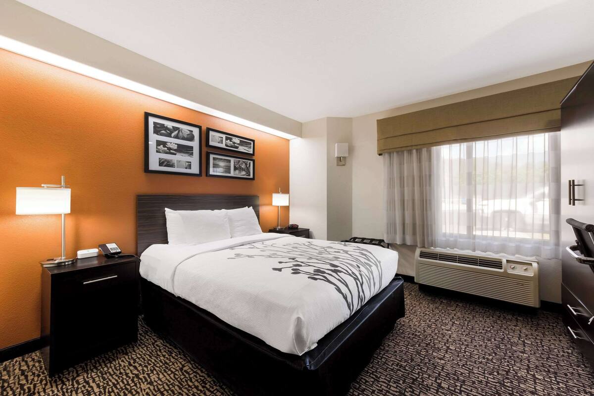 A hotel room with a bed, bedside tables, a lamp, a picture frame, a window with curtains, and an air conditioning unit.
