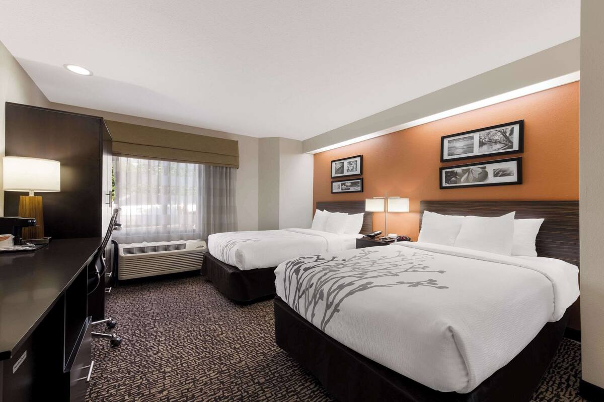 A hotel room with two beds, a desk, a chair, a lamp, and a window with curtains.
