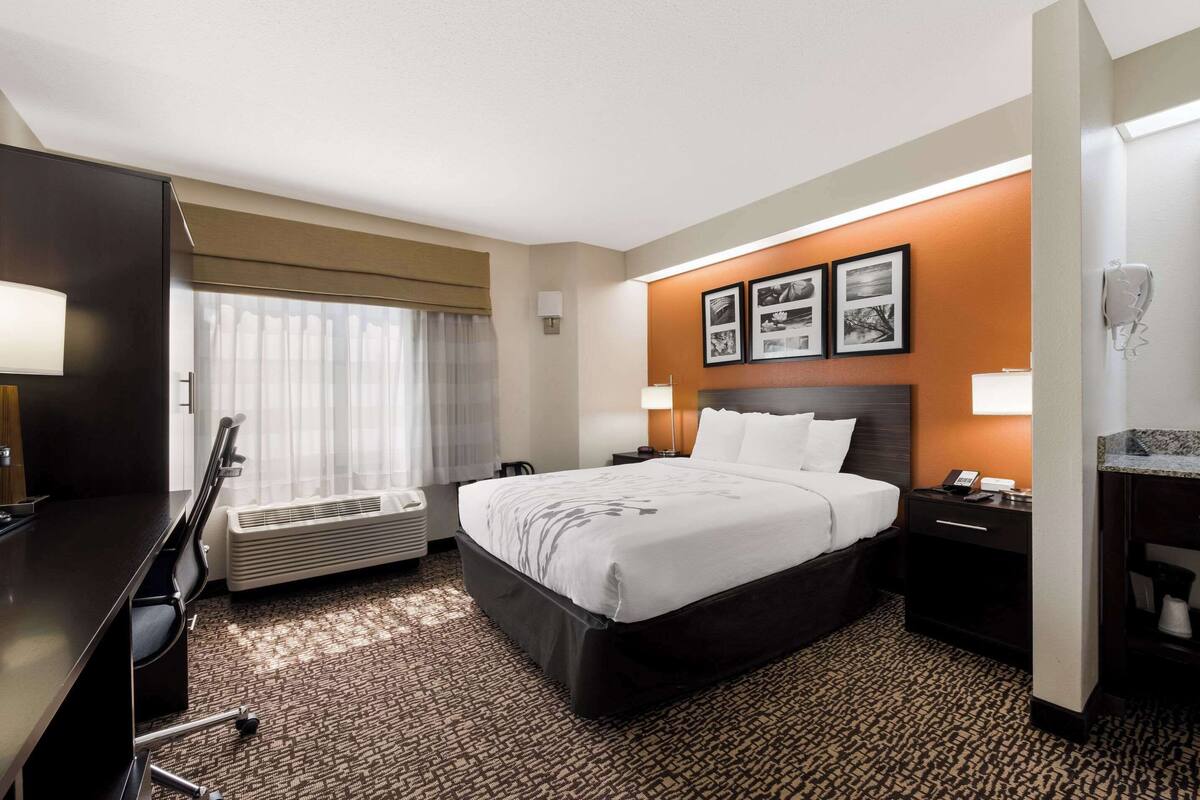 A hotel room with a large bed, a desk, a chair, an air conditioning unit, a nightstand with a phone, and framed pictures on the wall.