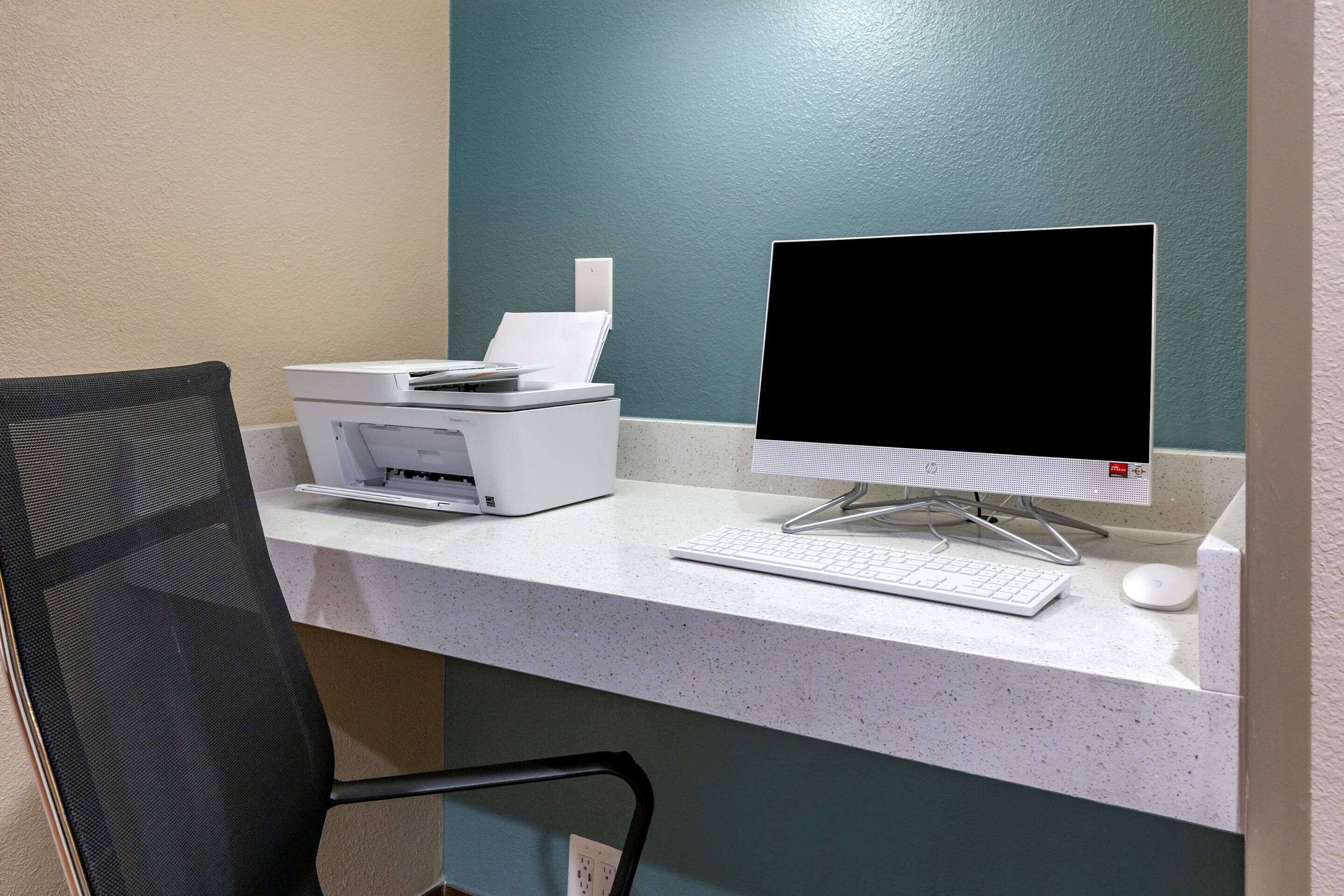 Un espacio de trabajo con una computadora de escritorio, una impresora y un teclado sobre un escritorio blanco frente a una pared verde azulado.