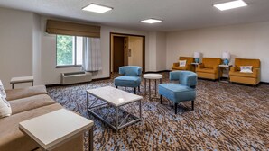 A hotel room with a beige sofa, a blue armchair, a small table, and a patterned carpet. There are two lamps on side tables and a door leading to another room. - Sleep Inn Fayetteville I-95 (Fayetteville)