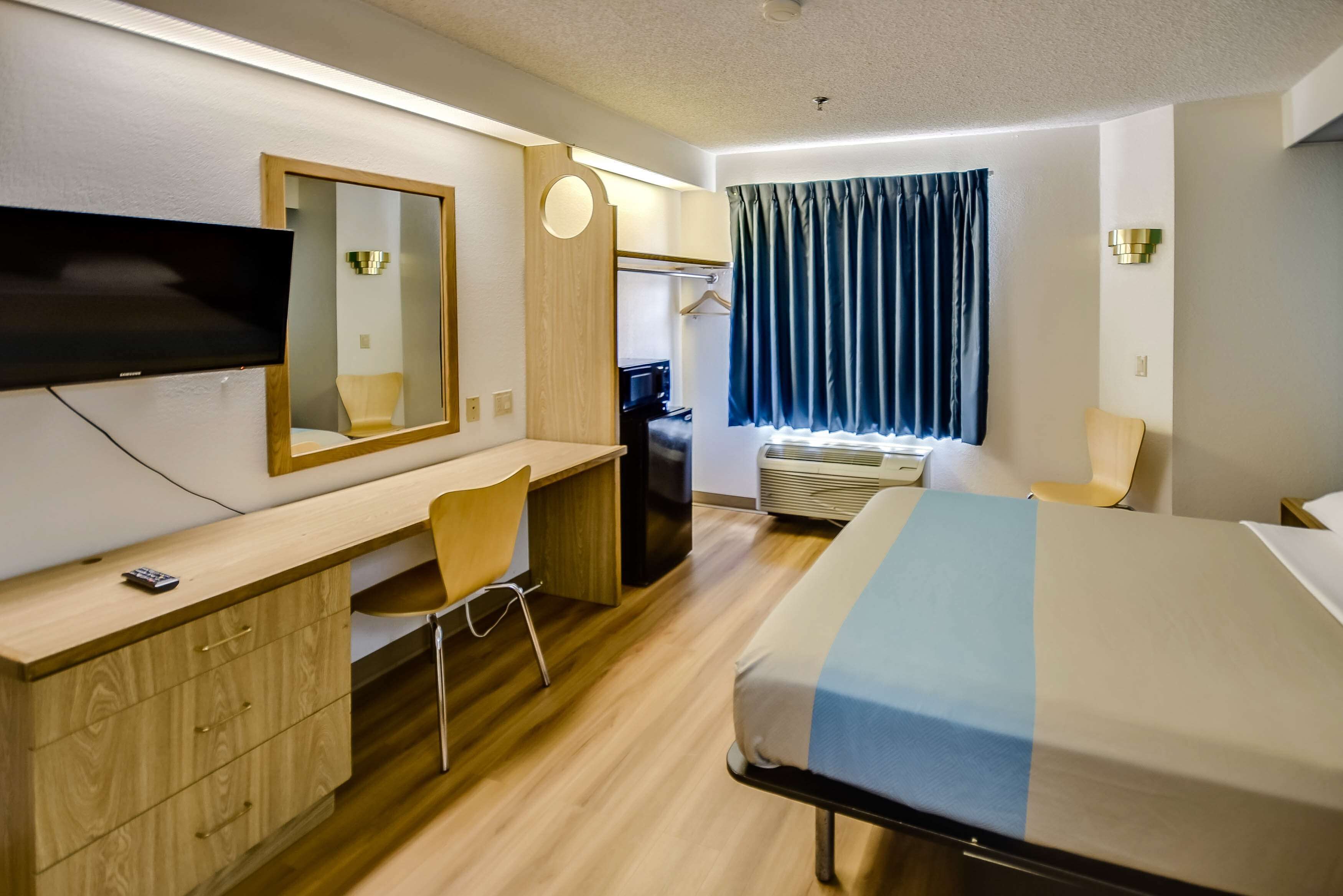 A hotel room with a bed, desk, chair, TV, mirror, and window with curtains.