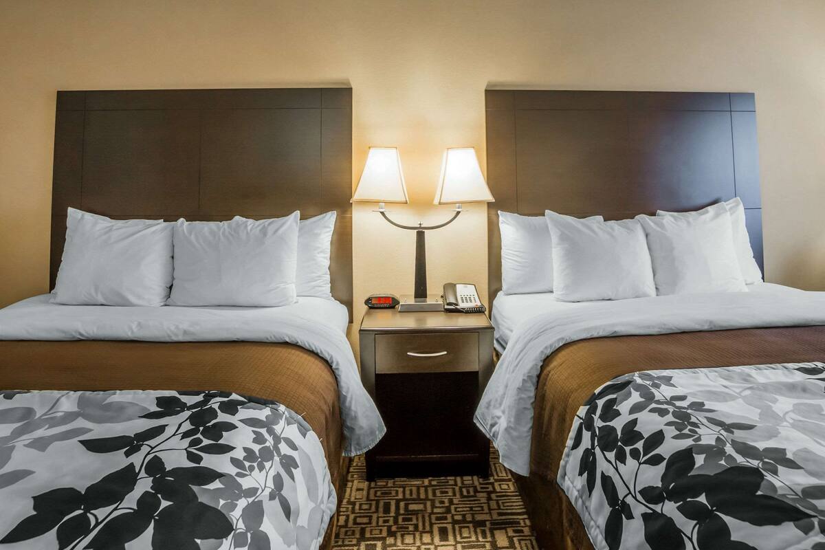 Habitación de hotel con dos camas, cada una con ropa blanca de cama y una cubierta de edredón con estampado floral. Hay una mesita de noche con una lámpara entre las camas, y un teléfono sobre la mesa.