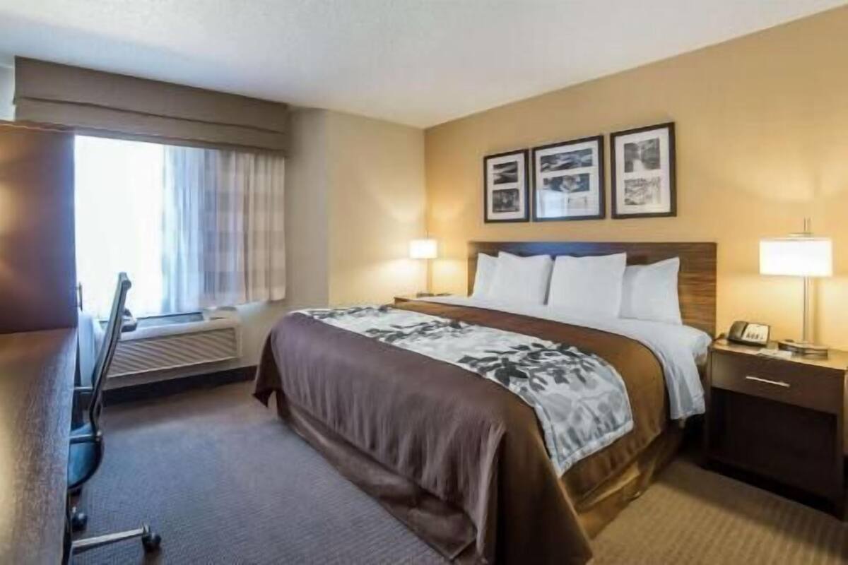 Una habitación de hotel con cama, mesitas de noche, escritorio, silla y ventana con cortinas.