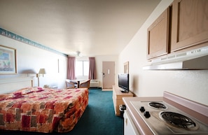 A hotel room with a bed, a desk with a TV, a kitchenette with a stove, and a window with curtains.