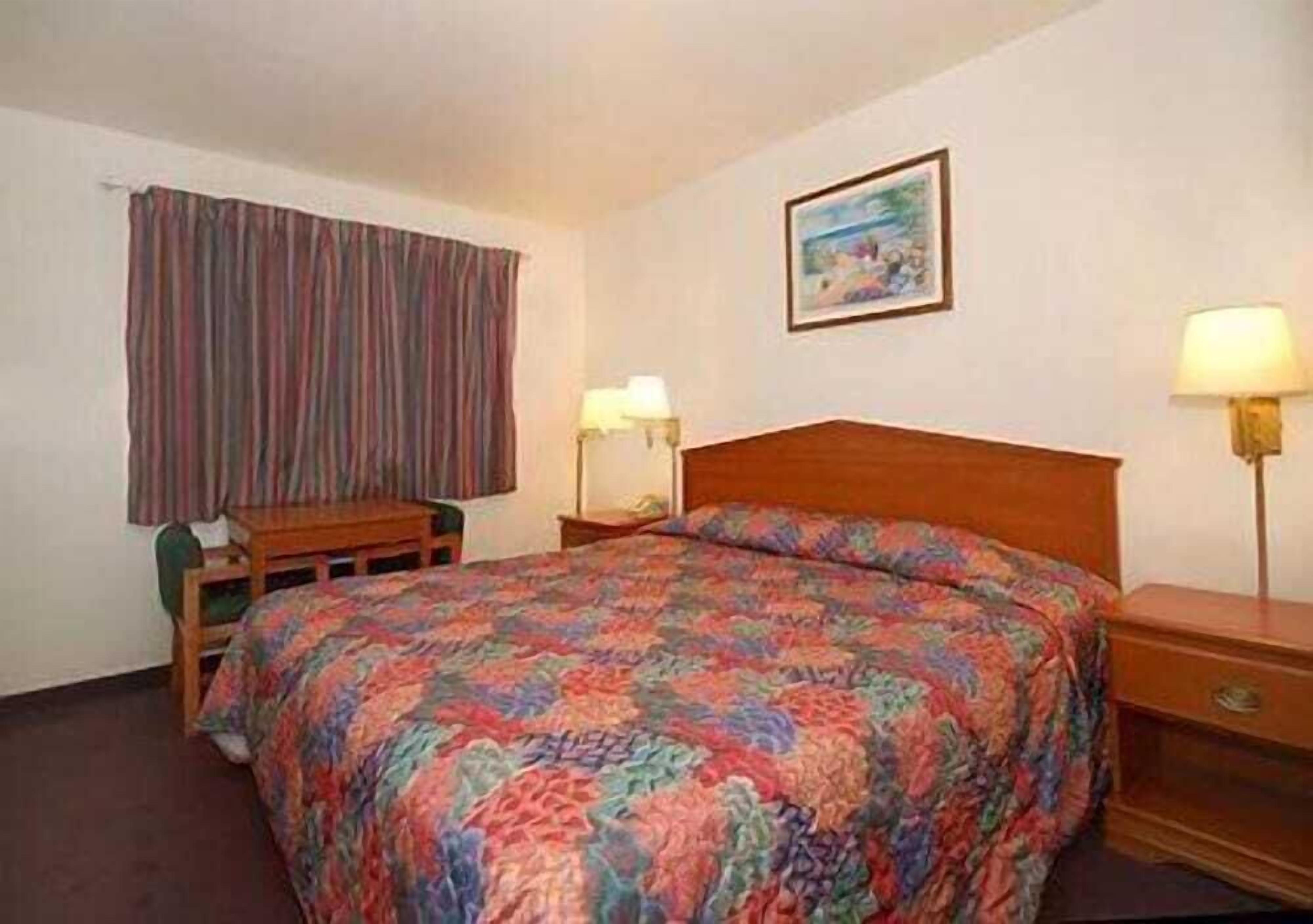 A hotel room with a bed, a desk, two bedside lamps, a window with curtains, and a painting on the wall.