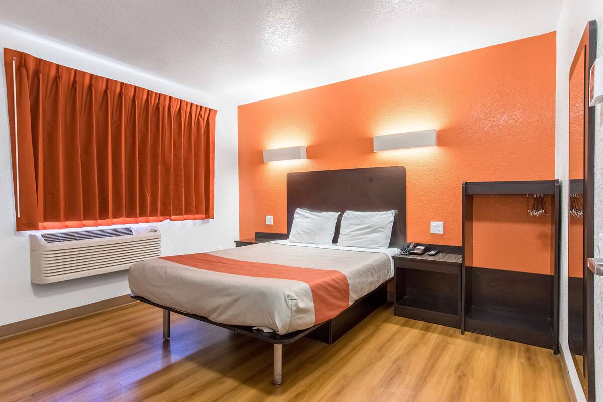 Quarto de hotel com cama, unidade de ar condicionado, mesa de cabeceira e luminária fixa na parede.