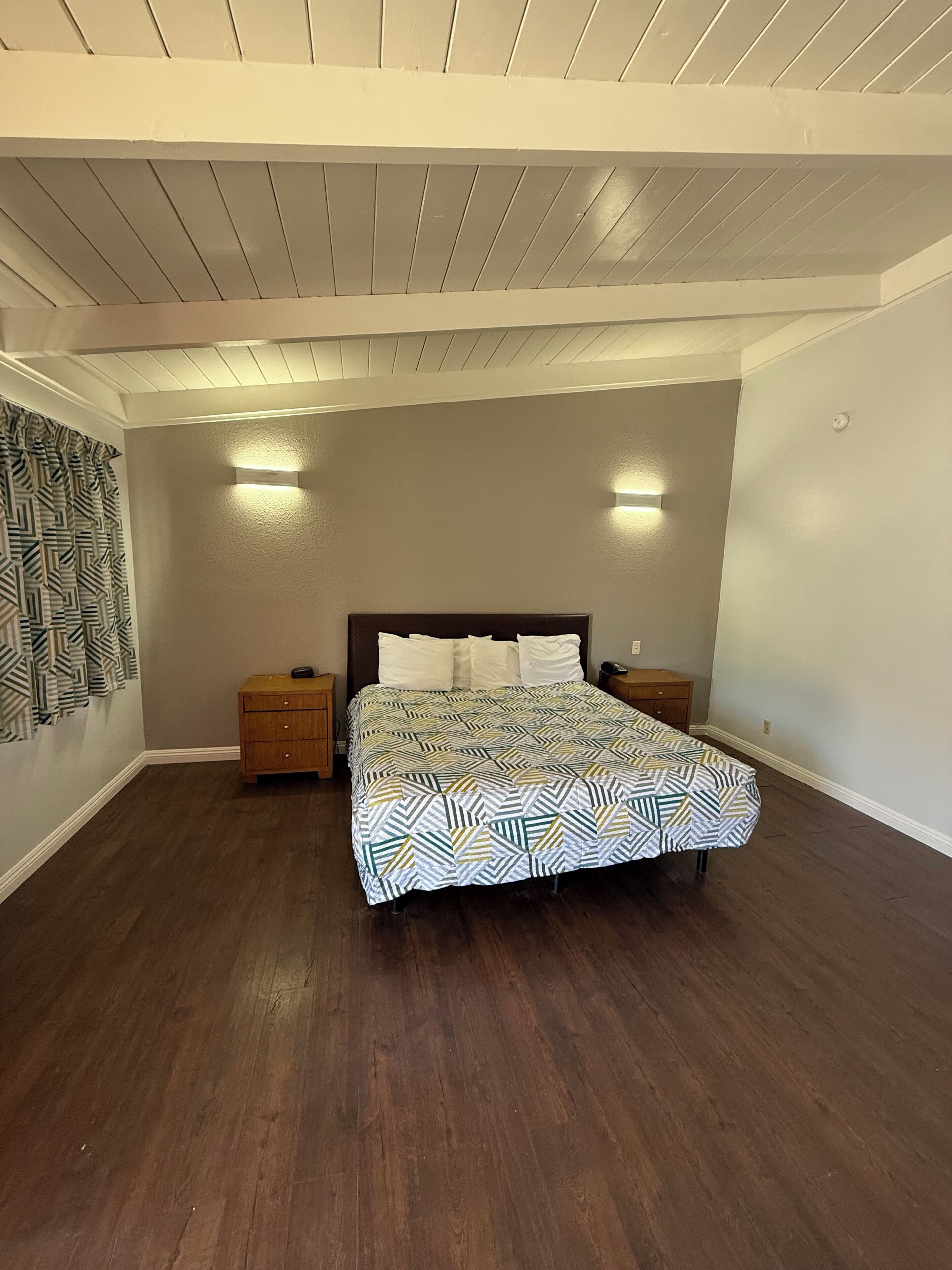 Un dormitorio con una cama, dos mesitas de noche, una colcha estampada, mesitas de noche de madera y un techo con vigas a la vista.