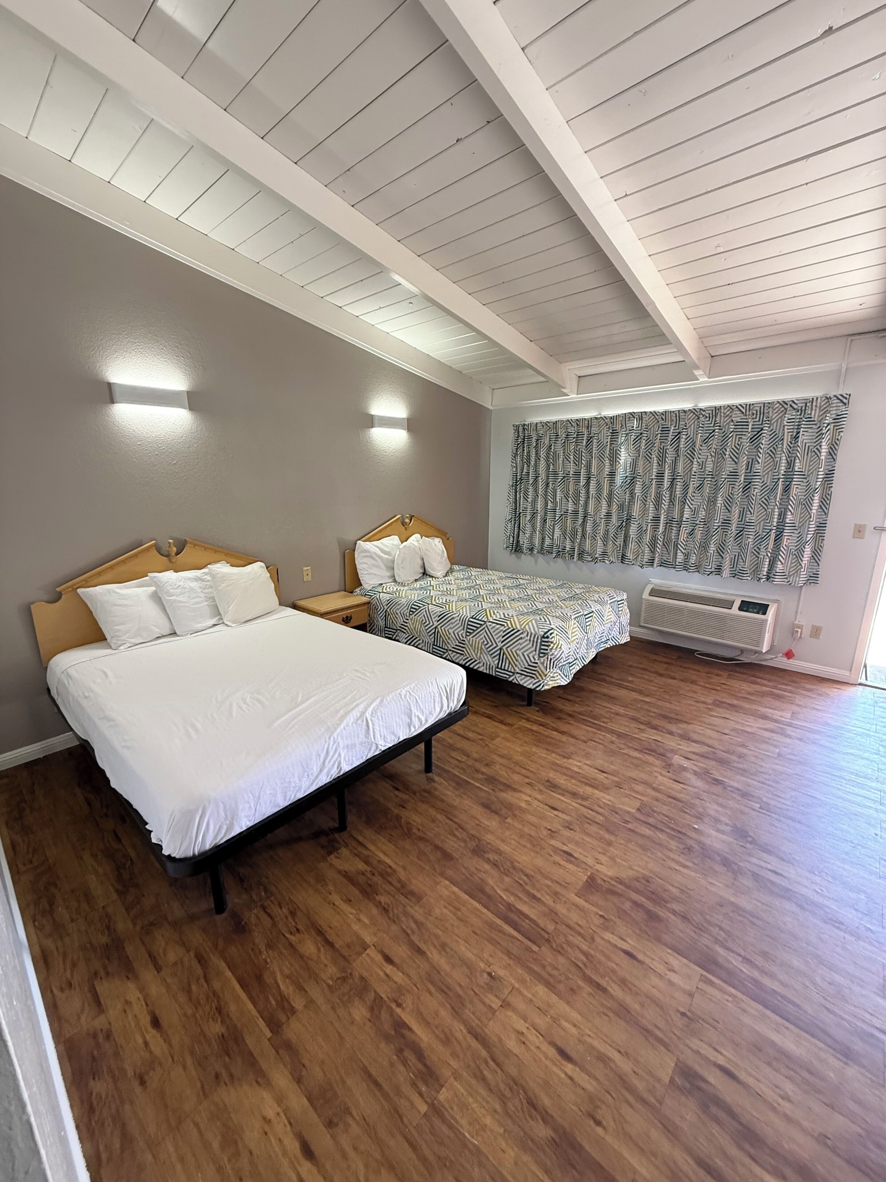 Un dormitorio con dos camas, suelo de madera, ventilador de techo y cortina estampada.