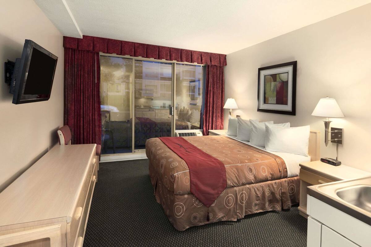 A hotel room with a bed, a desk, a TV, a window with curtains, a painting, and a lamp.