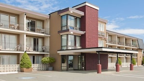 Un hotel de varios pisos con diseño moderno, que cuenta con balcones con mobiliario exterior, una entrada central con área cubierta y un estacionamiento.