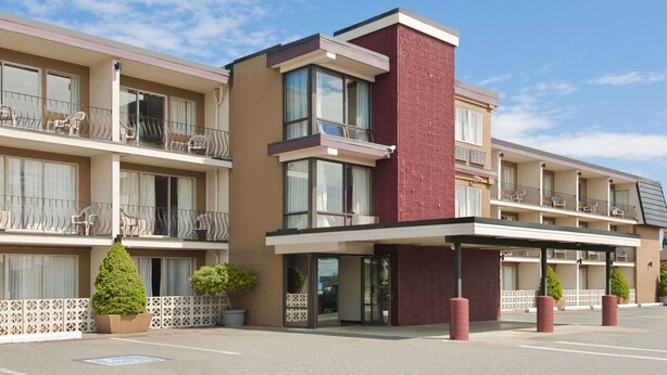 A multi-story hotel with a modern design, featuring balconies with outdoor furniture, a central entrance with a covered area, and a parking lot.