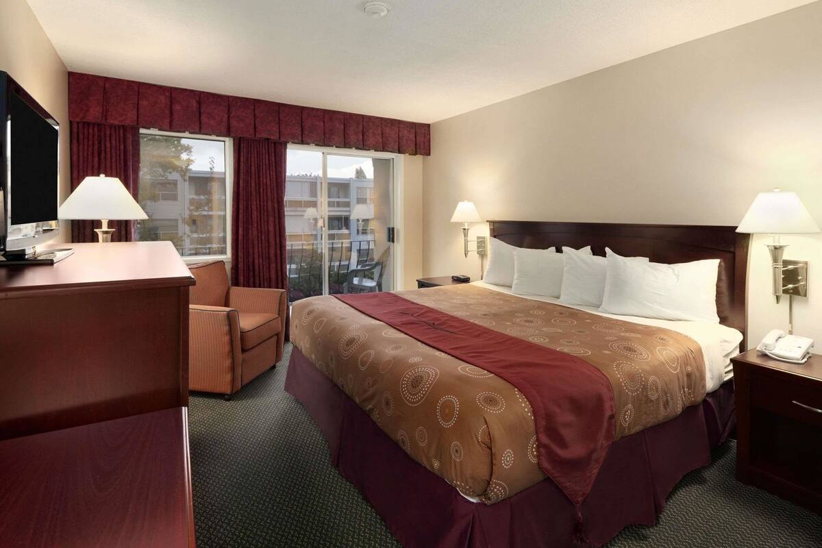 A hotel room with a bed, two bedside lamps, a desk with a computer, a chair, and a large window with curtains.