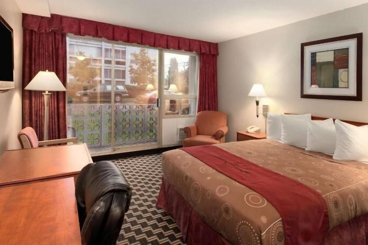 A hotel room with a bed, a desk, a chair, a window with curtains, and a painting on the wall.