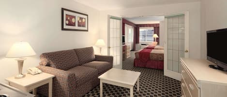 A hotel room with a patterned carpet, a sofa, a small table, a TV, and a kitchenette.
