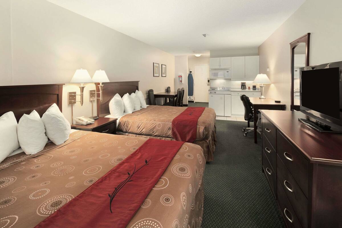 A hotel room with two beds, a desk, a chair, a television, and a kitchenette.