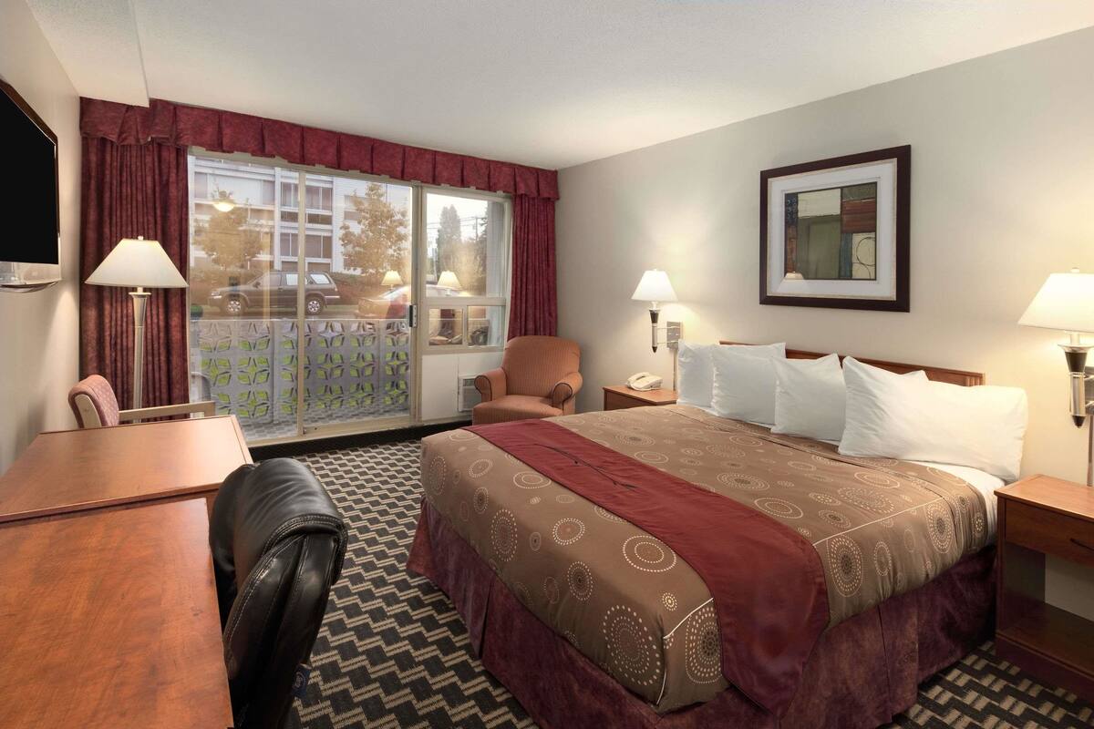 A hotel room with a large bed, a desk, a chair, a window with curtains, and a framed picture on the wall.