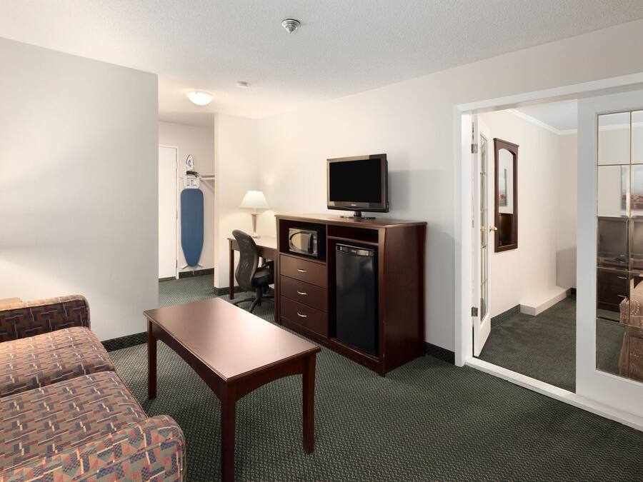 Ein Hotelzimmer mit einer Couch, einem Holztisch, einem Fernseher auf einem Schrank, einem Schreibtisch mit Stuhl und einer Glastür zu einem Balkon.