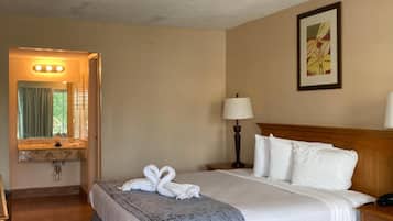A hotel room with a bed, a bedside table, a lamp, a framed picture on the wall, and a bathroom visible through an open door.