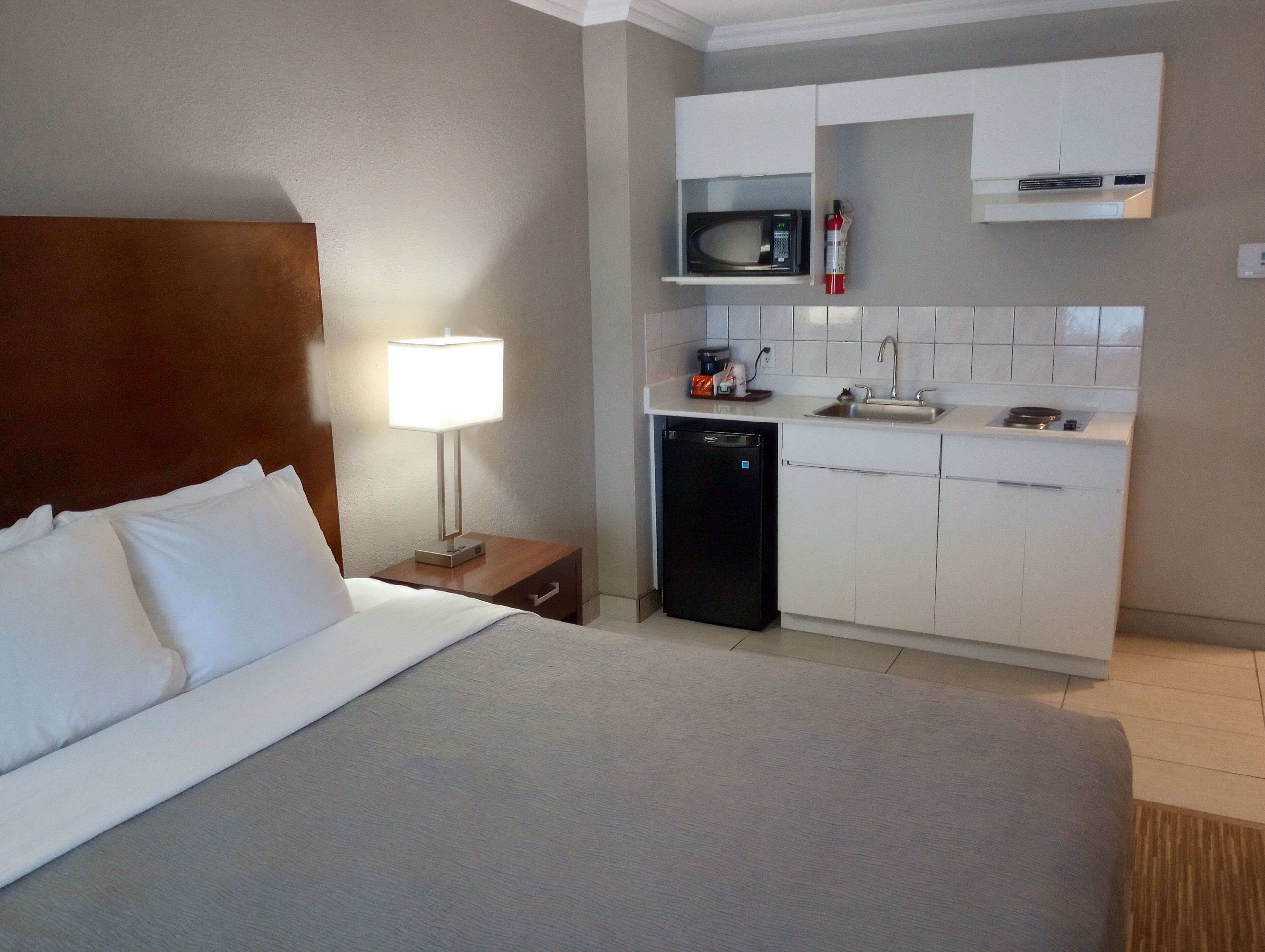A hotel room with a bed, a bedside lamp, a small kitchenette with a microwave, a sink, and a dishwasher.