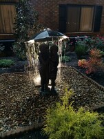 Un jardín con fuente de agua, que incluye una estatua y un paraguas, rodeado de guijarros y plantas.