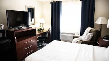 Habitación de hotel con cama, escritorio con televisor, silla, lámpara de escritorio, aire acondicionado y ventana con cortinas.