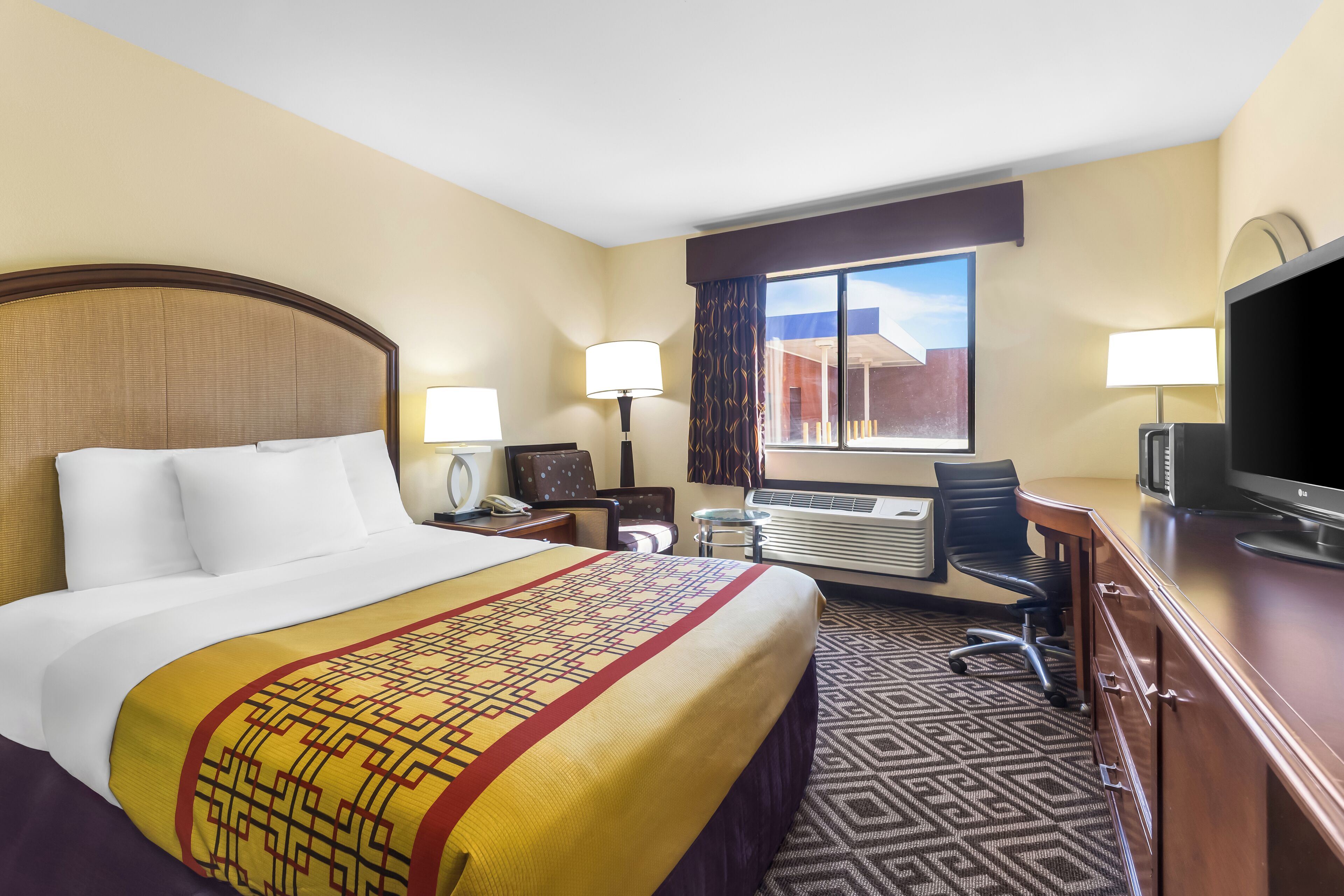 A hotel room with a large bed, a desk with a computer, a TV, and an air conditioning unit.