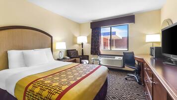 A hotel room with a large bed, a desk with a computer, a TV, and an air conditioning unit.