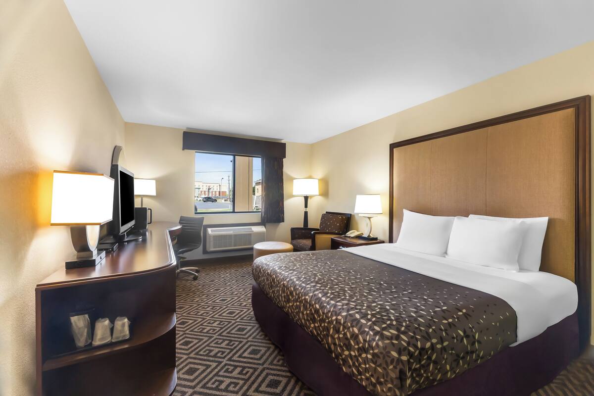 A hotel room with a large bed, a desk with a computer, a chair, a window with a view, and a wall-mounted TV.